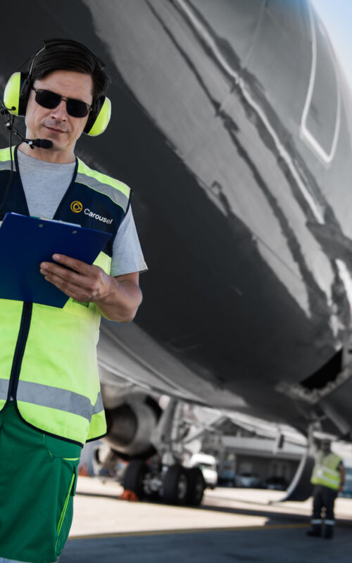 Active teamwork. Low angle portrait of serene man in sunglasses writing on clipboard. Colleagues, plane with open cargo door in the background