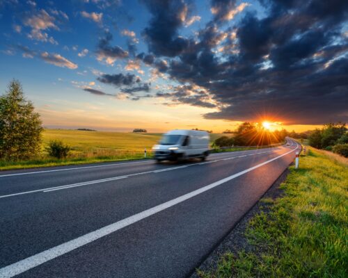 Motion blurred white fast delivery van driving on the asphalt road in rural landscape in the rays of the sunset with dark storm cloud A Carousel van travelling down the road on a delivery as the sun sets in the background