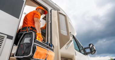 A Technician Wearing An Orange Shirt And Carrying A Tool Bag Enters An RV. He Is Wearing Safety Glasses And A Cap. The RV Is White And Has A Large Side Door. The Image Is Taken From A Low Angle, Looking Up At The Technician.